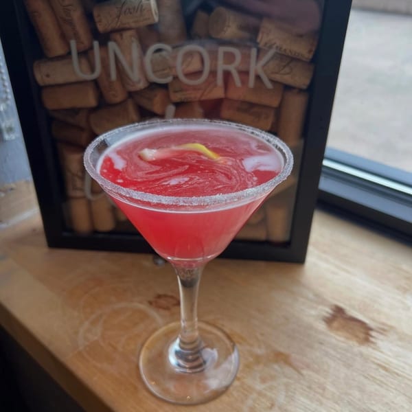 A sugar-rimmed red martini with a lemon twist on a bar top in front of an Uncork wine cork display.
