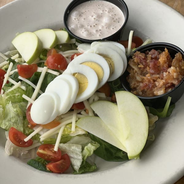 A fresh salad with lettuce, tomatoes, cheese, sliced hard-boiled eggs, green apple, bacon bits, and dressing.