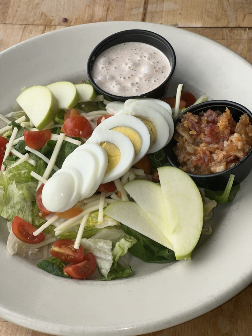 A fresh salad with lettuce, tomatoes, cheese, sliced hard-boiled eggs, green apple, bacon bits, and dressing.